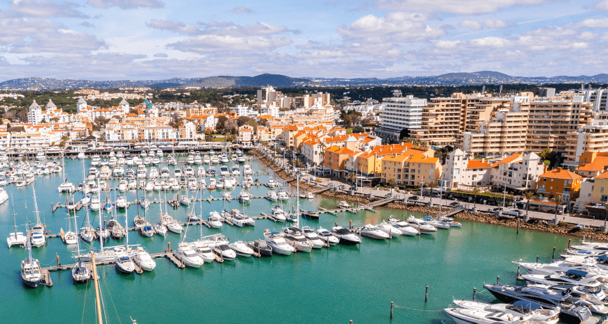 Luxury yachts and sleek modern speedboats moored up alongside the scenic boardwalk restaurants at the vibrant Vilamoura Marina under a clear blue sky